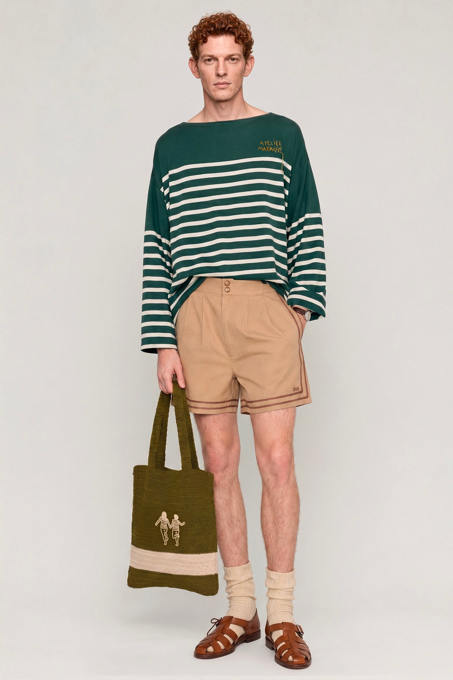 Man wearing a green and white Breton shirt by Atelier Marinière with beige tailored shorts and a knitted tote bag, handcrafted in Italy from premium cotton.