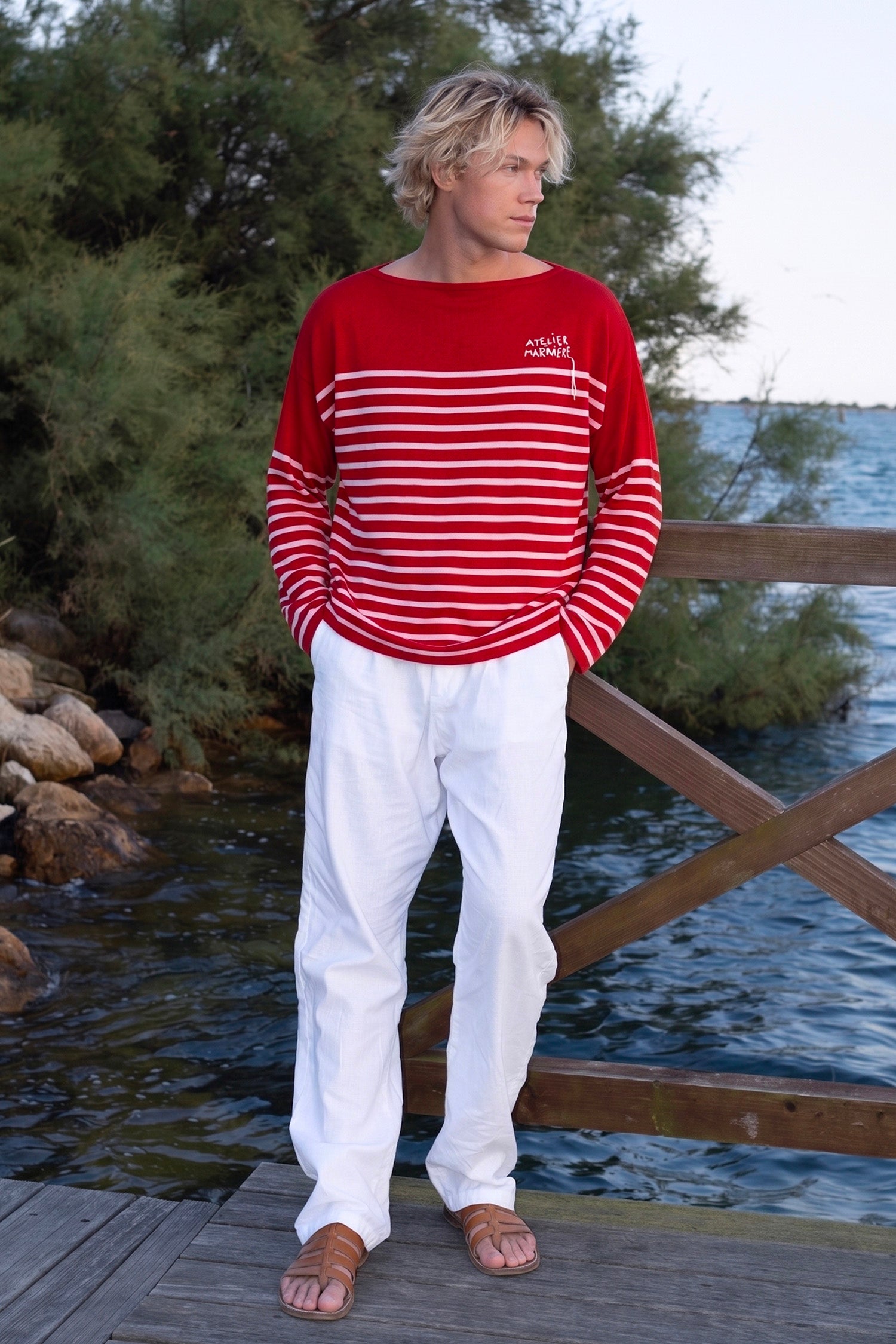 Model wearing Atelier Marinière's oversized red Breton shirt on a waterfront promenade in the Venetian lagoon, handcrafted in Italy from premium cotton.